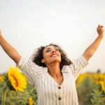 A beautiful woman raising her hands in the air like a champion because she is happy for overcoming negative thoughts in a field of sunflowers.