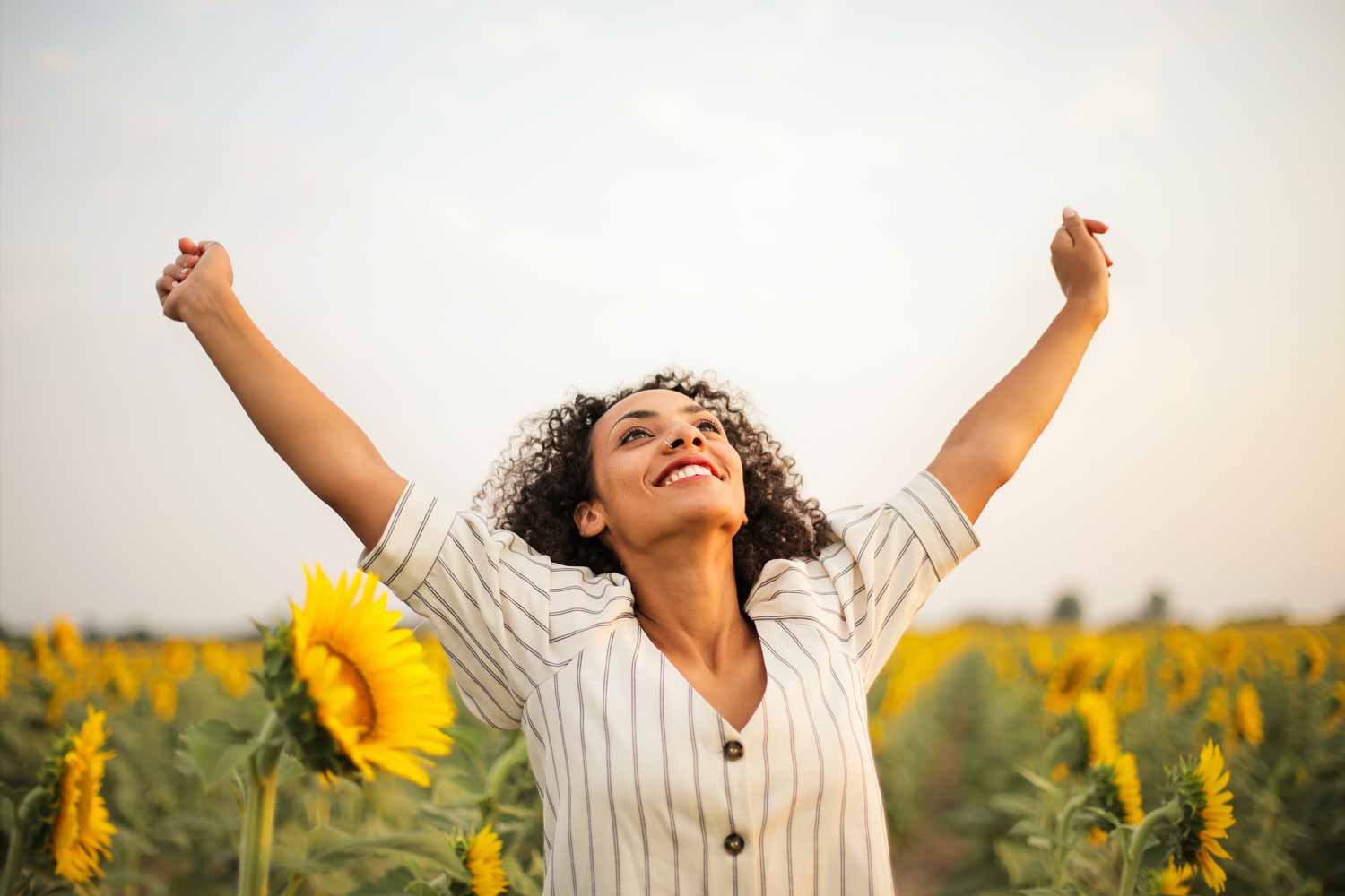 A beautiful woman raising her hands in the air like a champion because she is happy for overcoming negative thoughts in a field of sunflowers.